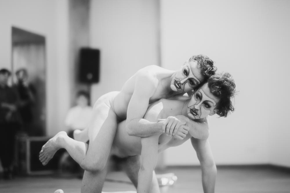A nude person rests on another nude person's back, the second person is on hands and knees