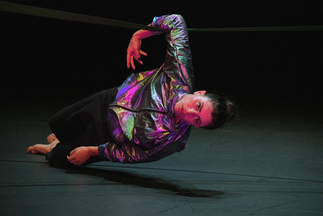 A dancer in a holographic jacket hanging onto a rope with one arm
