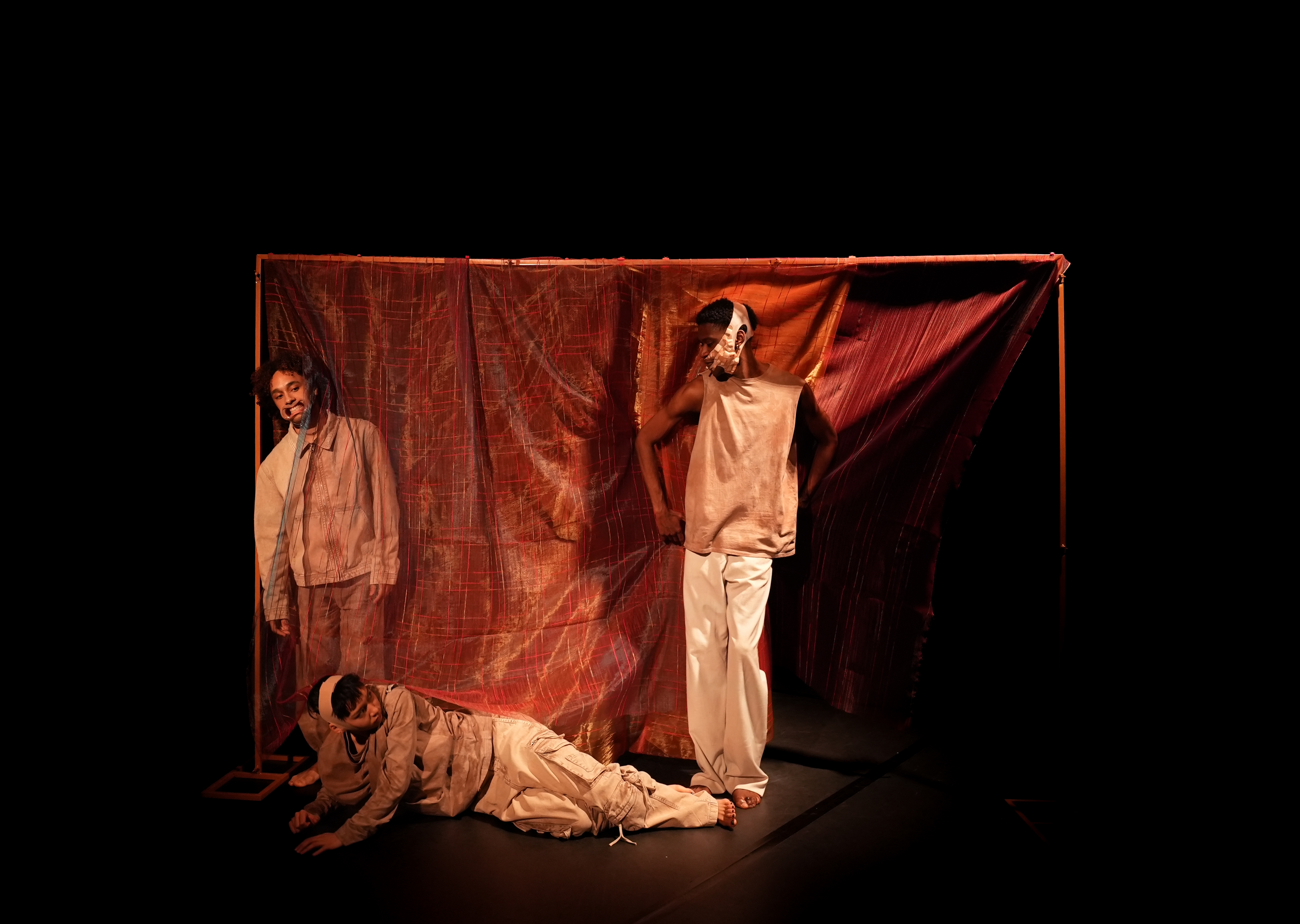Three performers, two standing, one lying on the floor, in front of a red sheet