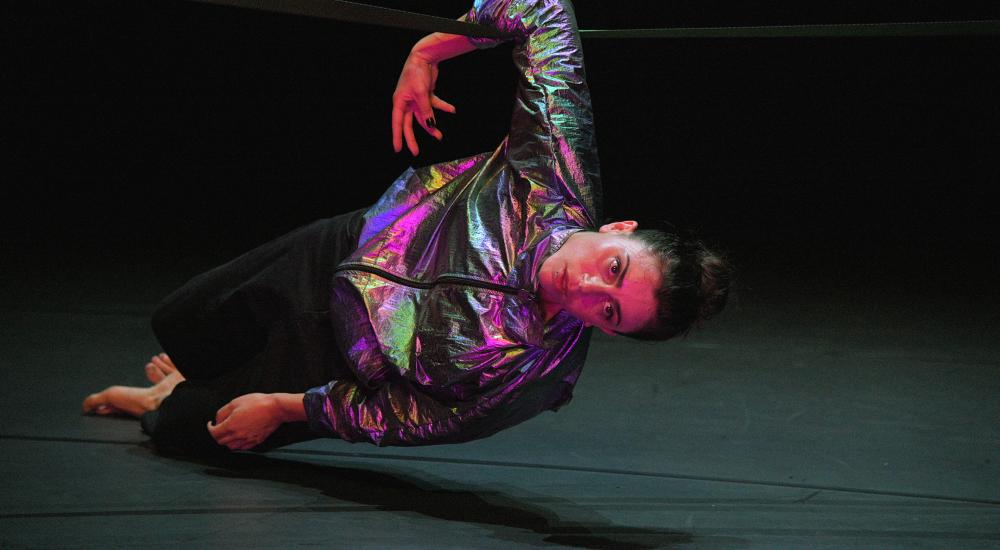 A dancer in a holographic jacket hanging onto a rope with one arm