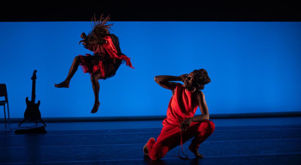Two Black dancers, one kneeling, one jumping in the air, wearing red in front of a blue screen