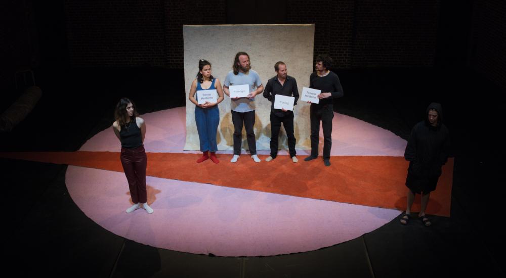 5 actors standing in a row on stage, four of them with a sheet in their hands on which their role is written