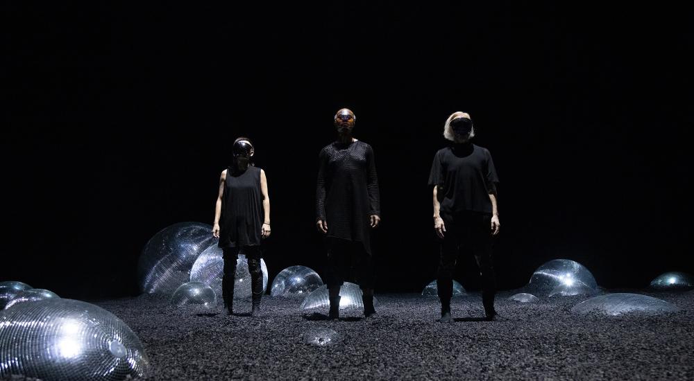 Mieko, Omagbitse and Meg wearing black masks standing in a desolate landscape littered with half-buried disco balls