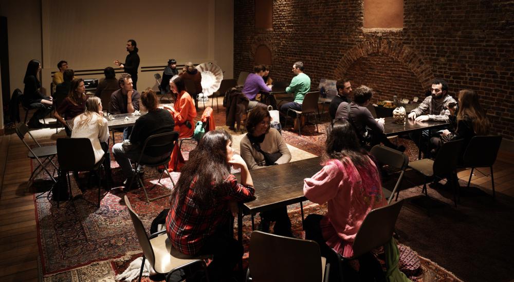 Around 20 people sitting at tables talking, with Persian rugs on the floor