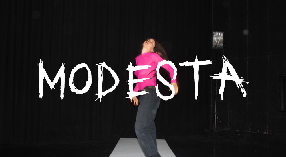 A young woman in a pink sweater standing on a white sheet with the word Modesta on top of it 
