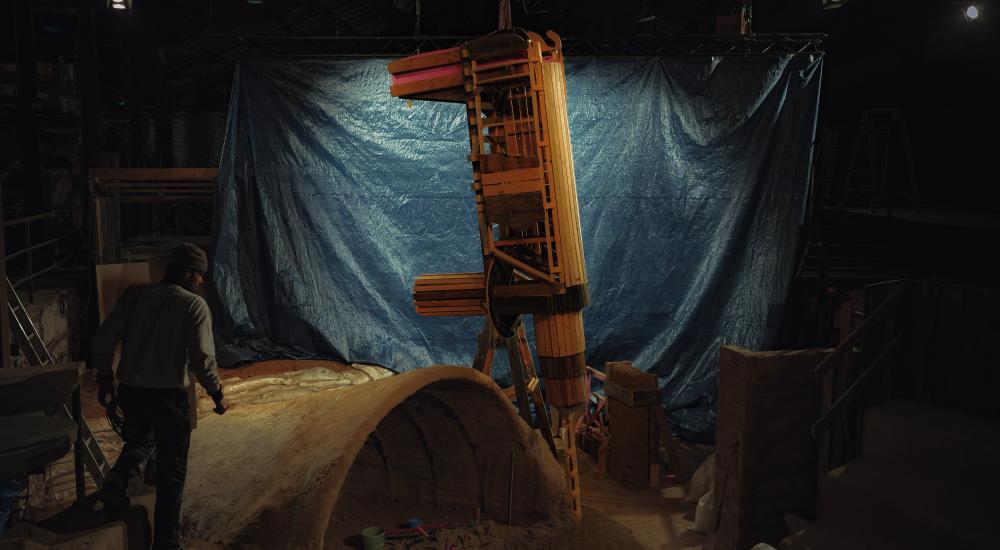 een houten constructie wordt omhoog getakeld in een donkere ruimte