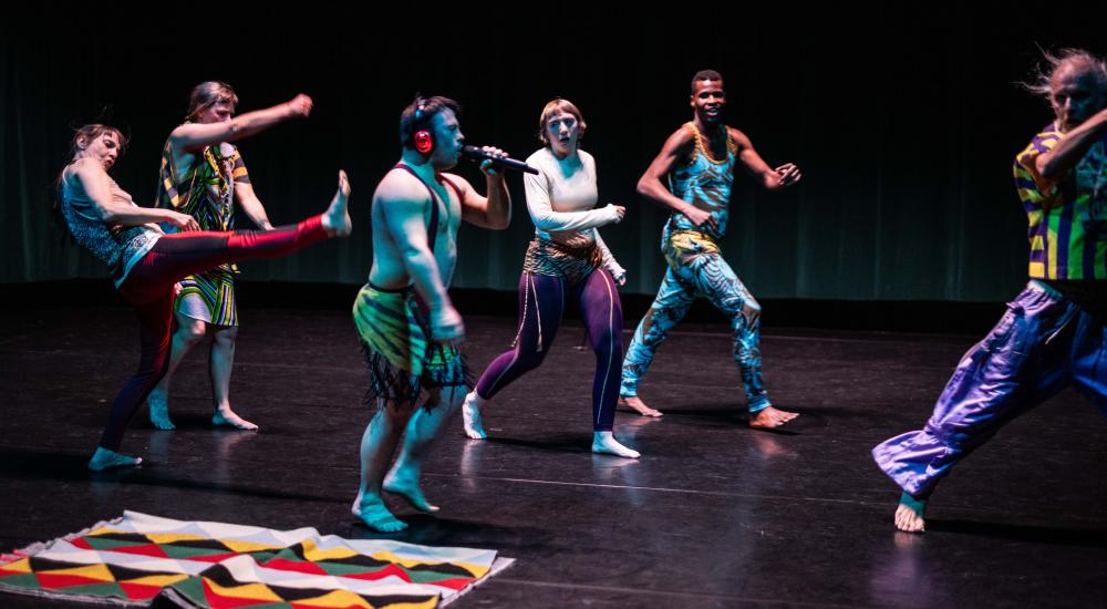 6 dancers wearing colourful outfits, one holding a mic and wearing headphones