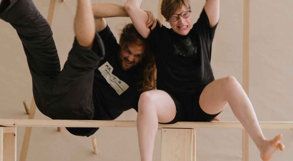 Twee personen die samen op een houten bank zitten. Ze zijn gekleed in zwarte shirts en shorts of broeken. Hun houding is dynamisch en speels: één persoon leunt achterover met de benen omhoog, terwijl de ander rechtop zit met de armen in de lucht. De setting is minimalistisch, met een lichte vloer en een eenvoudige achtergrond van houten panelen.