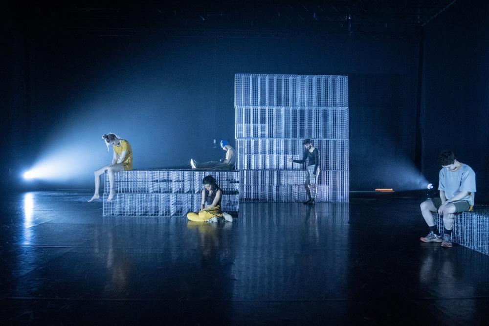 Five performers scattered over the stage sitting in front of a big metal construction