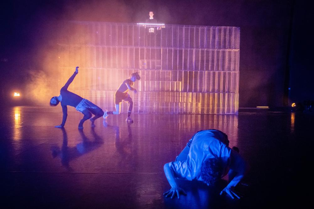 3 performers moving in front of a big metal construction that has one person standing on it