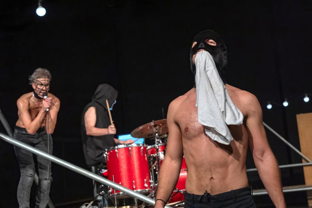 One person wearing a mask holding a cloth in their mouth in front of someone singing into a mic and someone else playing drums