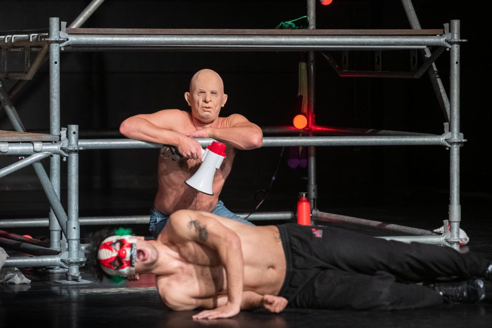 Two dancers wearing masks, one lying on the floor, one sitting behind him holding a megaphone