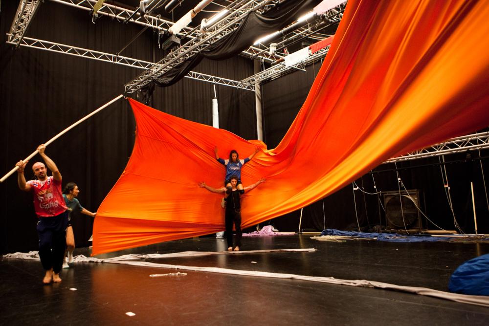 A performer sitting on another's shoulders in front of an orange sheet held up by another performer on the left