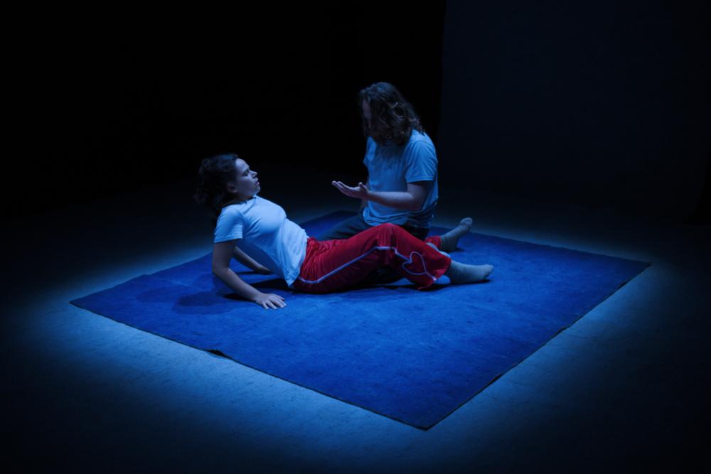 two actors in conversation, one lying on the ground with torso thrust upwards, the other on crouch