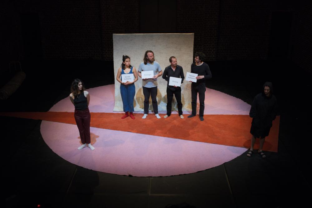 5 actors standing in a row on stage, four of them with a sheet in their hands on which their role is written