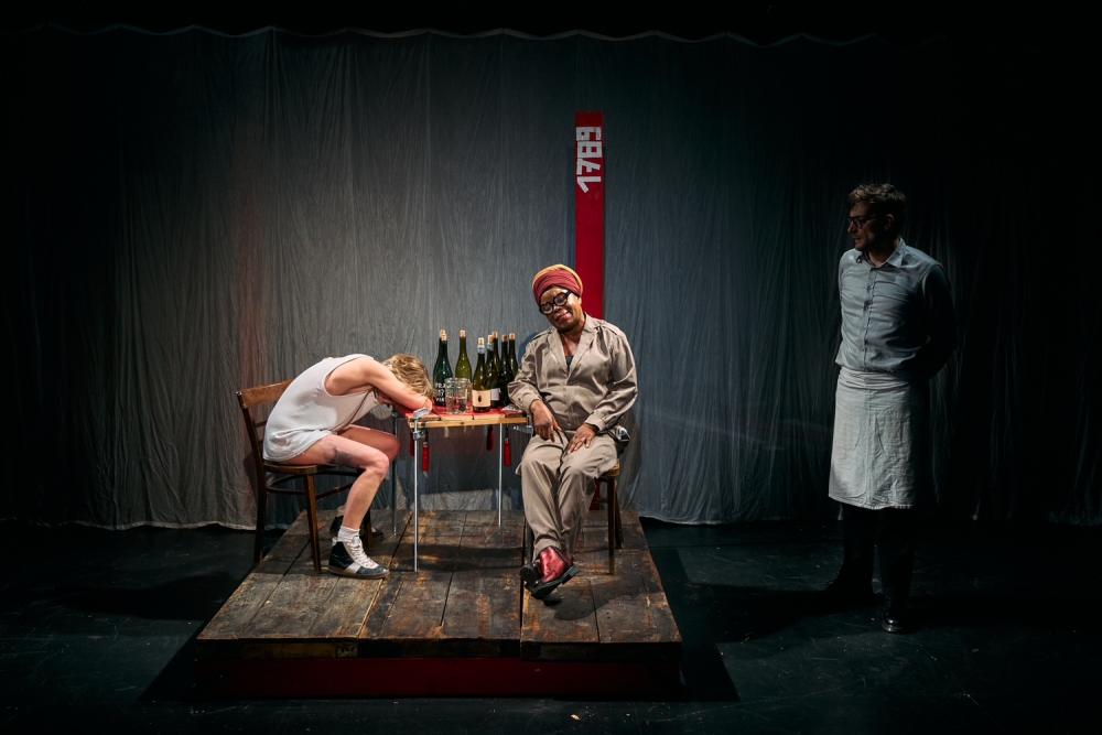 Aurélie and Starlette sitting at a table, Aurélie passed out and Michiel standing to the side, dressed as a waiter