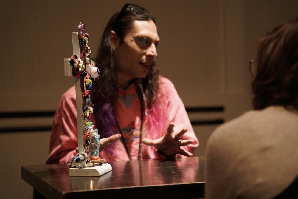 A person with long hair sitting at a table next to a bedazzled cross