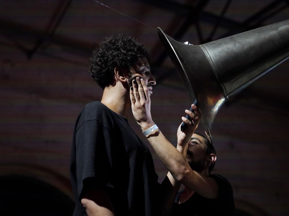person standing under a huge horn reading its smartphone while another person standing right of him is holding his face with one hand