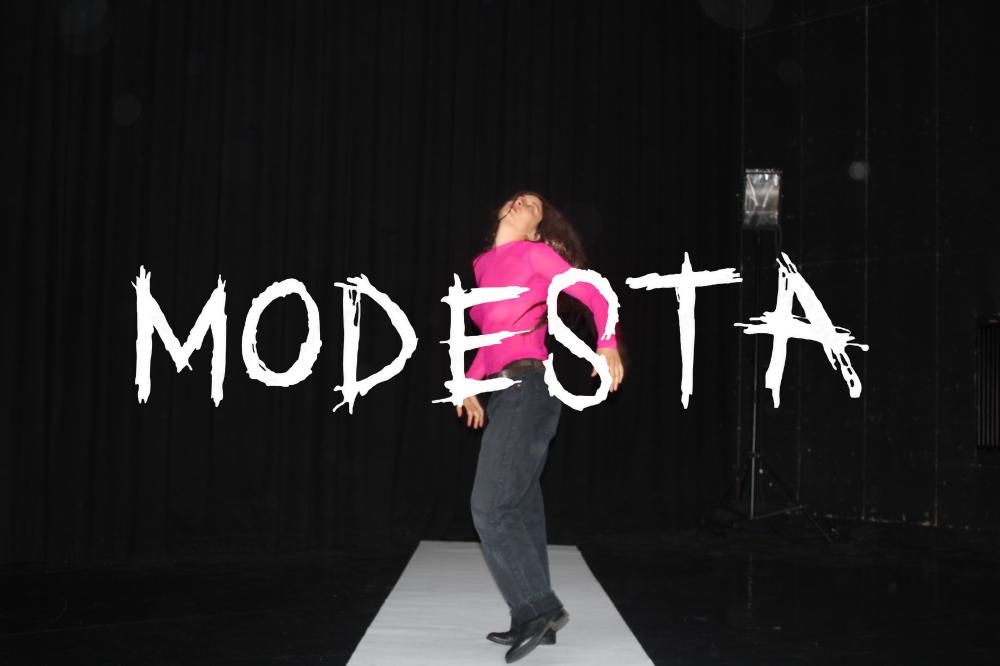 A young woman in a pink sweater standing on a white sheet with the word Modesta on top of it 