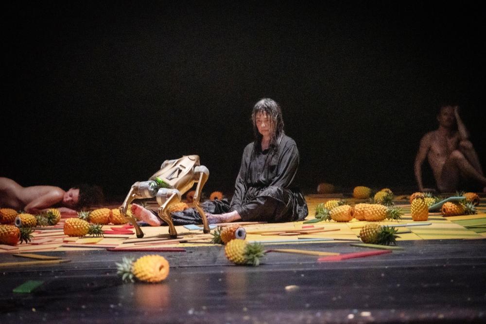 A person sitting on the floor between fruit with a robot dog in the foreground and two people lying and sitting naked in the background