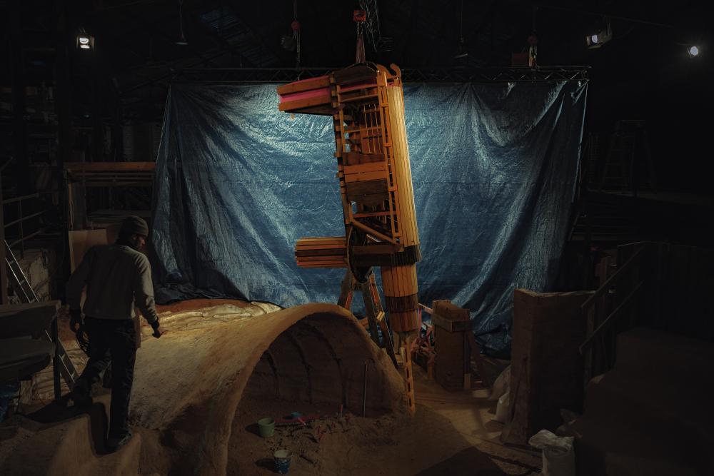 een houten constructie wordt omhoog getakeld in een donkere ruimte