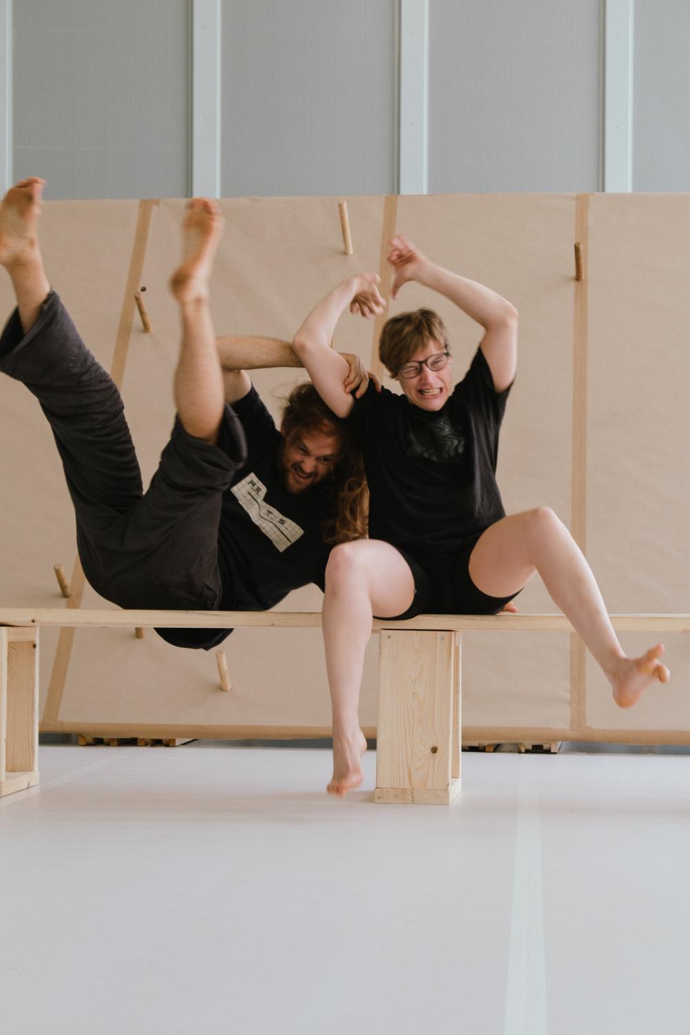 Twee personen die samen op een houten bank zitten. Ze zijn gekleed in zwarte shirts en shorts of broeken. Hun houding is dynamisch en speels: één persoon leunt achterover met de benen omhoog, terwijl de ander rechtop zit met de armen in de lucht. De setting is minimalistisch, met een lichte vloer en een eenvoudige achtergrond van houten panelen.
