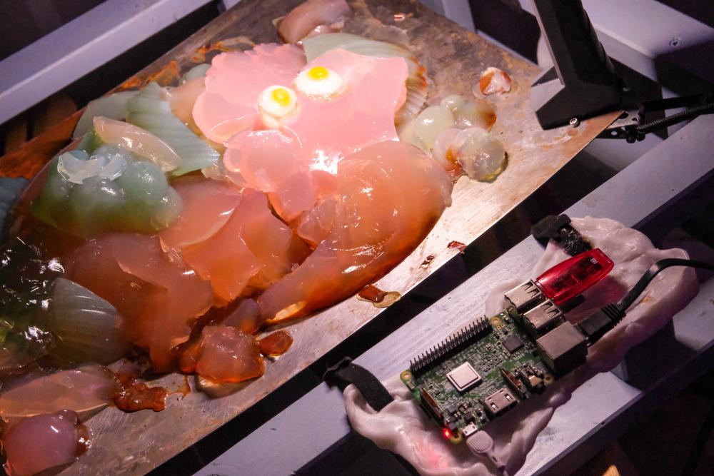 A close-up of an artistic or experimental setup. On a metallic surface, there are various translucent, jelly-like blobs in different colors (pink, green, orange, and clear), some of which are illuminated from below, giving a glowing effect.