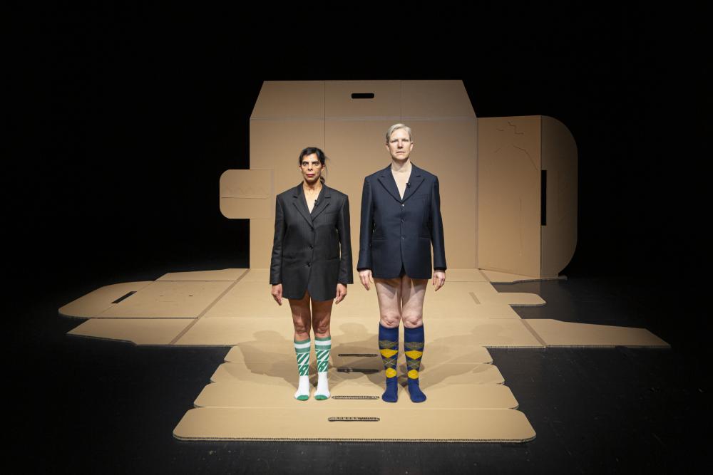 Two people wearing a suit jacket and no trousers standing on a huge unfolded cardboard box