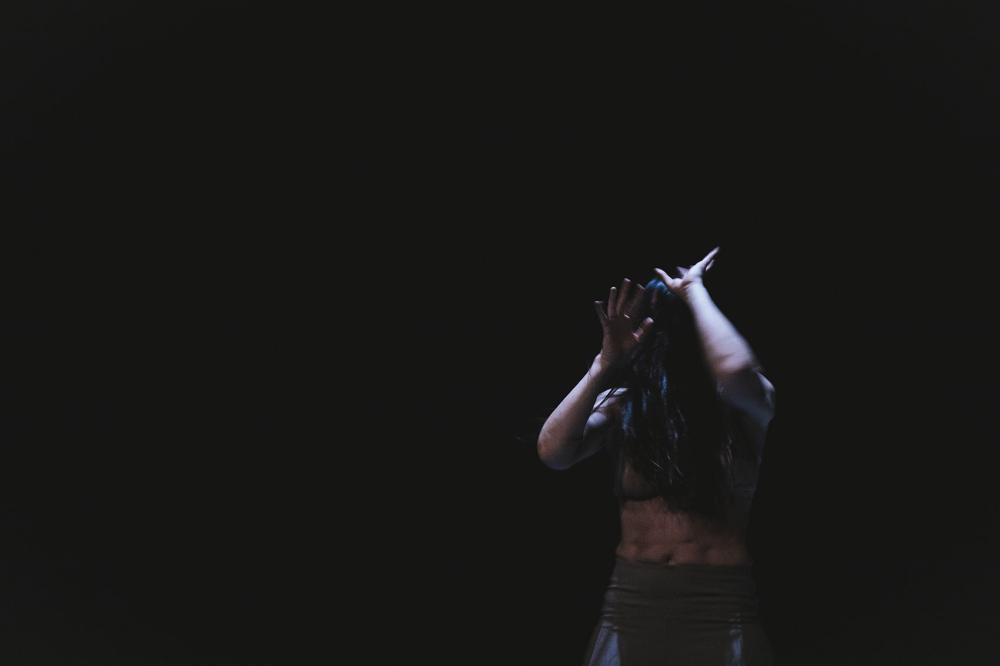 A dancer in front of a dark background holding their arms up in front of their face