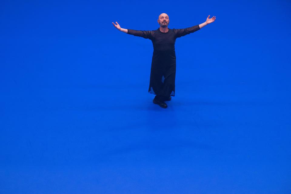 1 performer with black clothes on a bright blue floor, spreading both arms and looking up to the sky