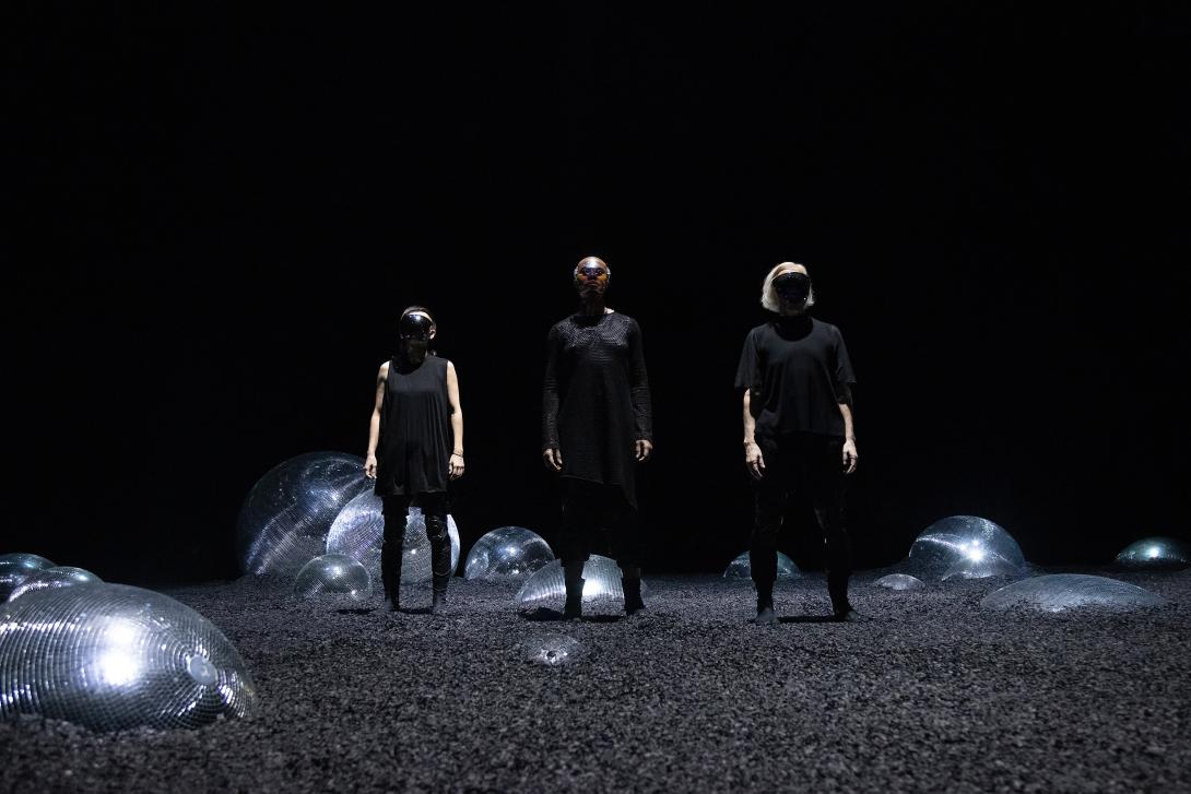 Mieko, Omagbitse and Meg wearing black masks standing in a desolate landscape littered with half-buried disco balls