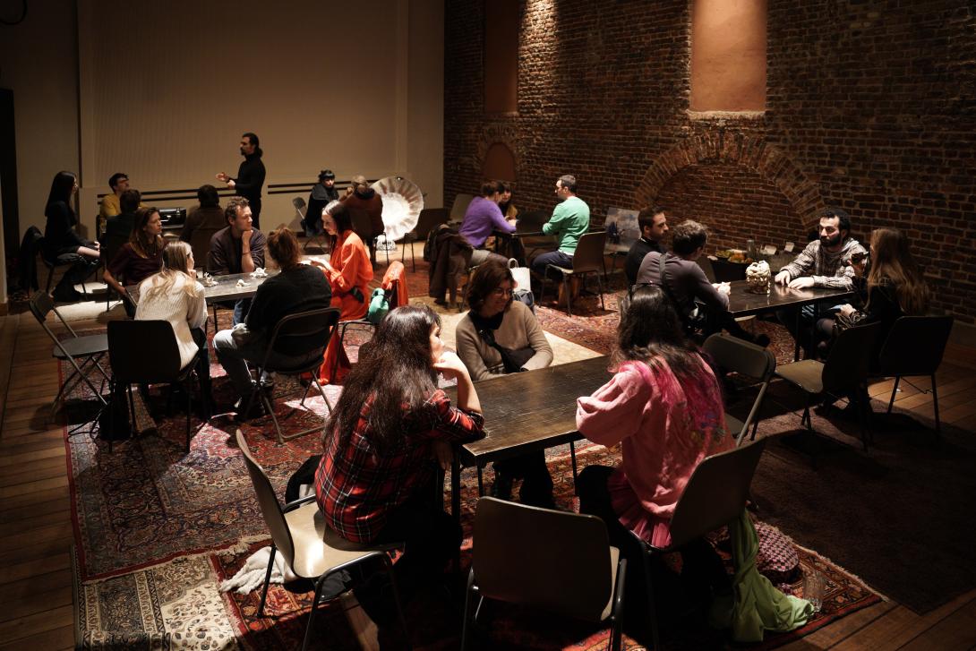 Around 20 people sitting at tables talking, with Persian rugs on the floor